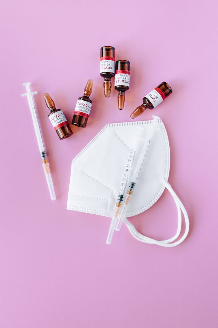 Flat lay of COVID-19 vaccine vials, syringes, and a face mask on a pink background.