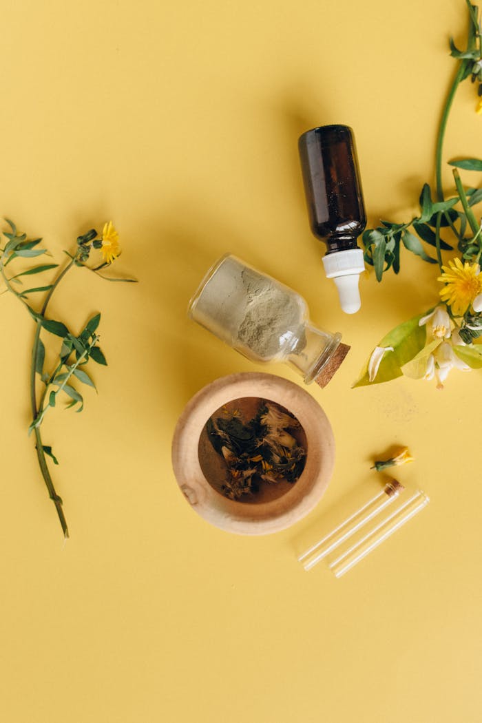 Flat lay of herbal medicine ingredients with glass bottles and leaves on yellow surface.